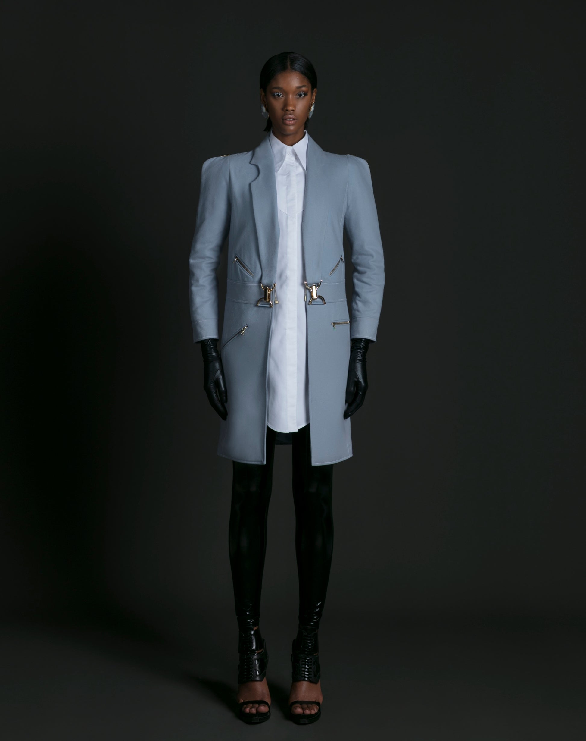 Person wearing a light gray coat, white shirt, black pants, and gloves on a dark background